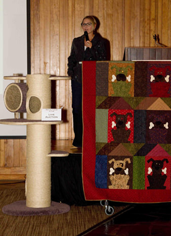 Emcee Kim Maus (from Good Day Oregon) showcasing two of the live auction items, Royal Meow cat castle and hand-made dog quilt.